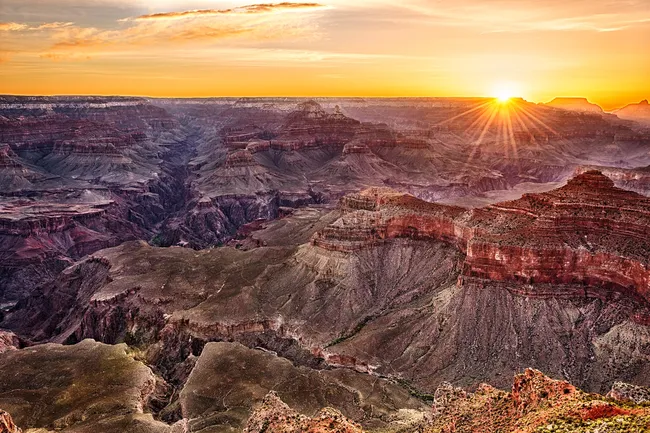 A breathtaking sunset over Yavapai Point at the Grand Canyon, with vibrant orange and yellow light rays reflecting off the deep canyon walls and plateaus.