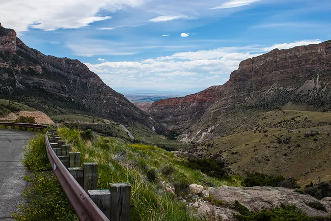 A winding road with wooden guardrails curves through the rugged cliffs of Bighorn Canyon under a bright blue sky.