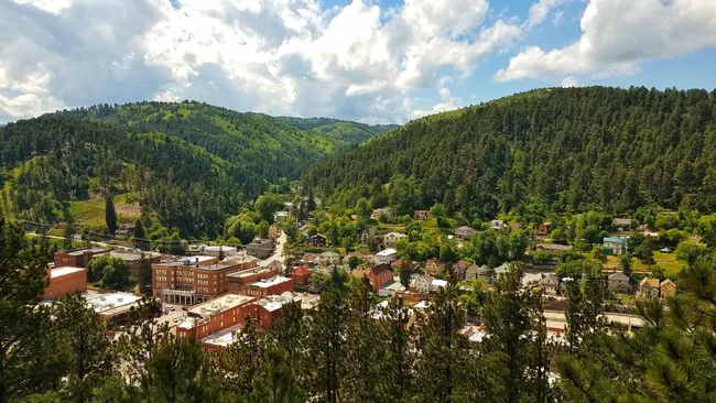 Aerial view of Deadwood, South Dakota, surrounded by forested green hills under a partly cloudy sky.