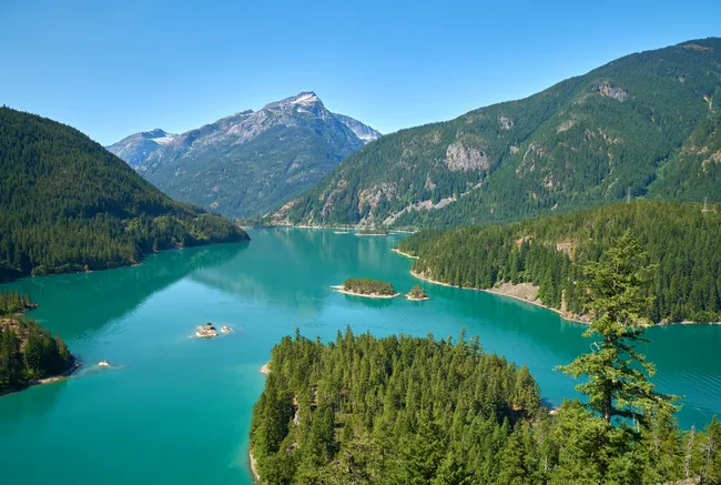 Alt text: Panoramic view of a turquoise mountain lake winding between forested islands and evergreen-covered slopes, with rugged peaks rising in the distance under a clear blue sky.