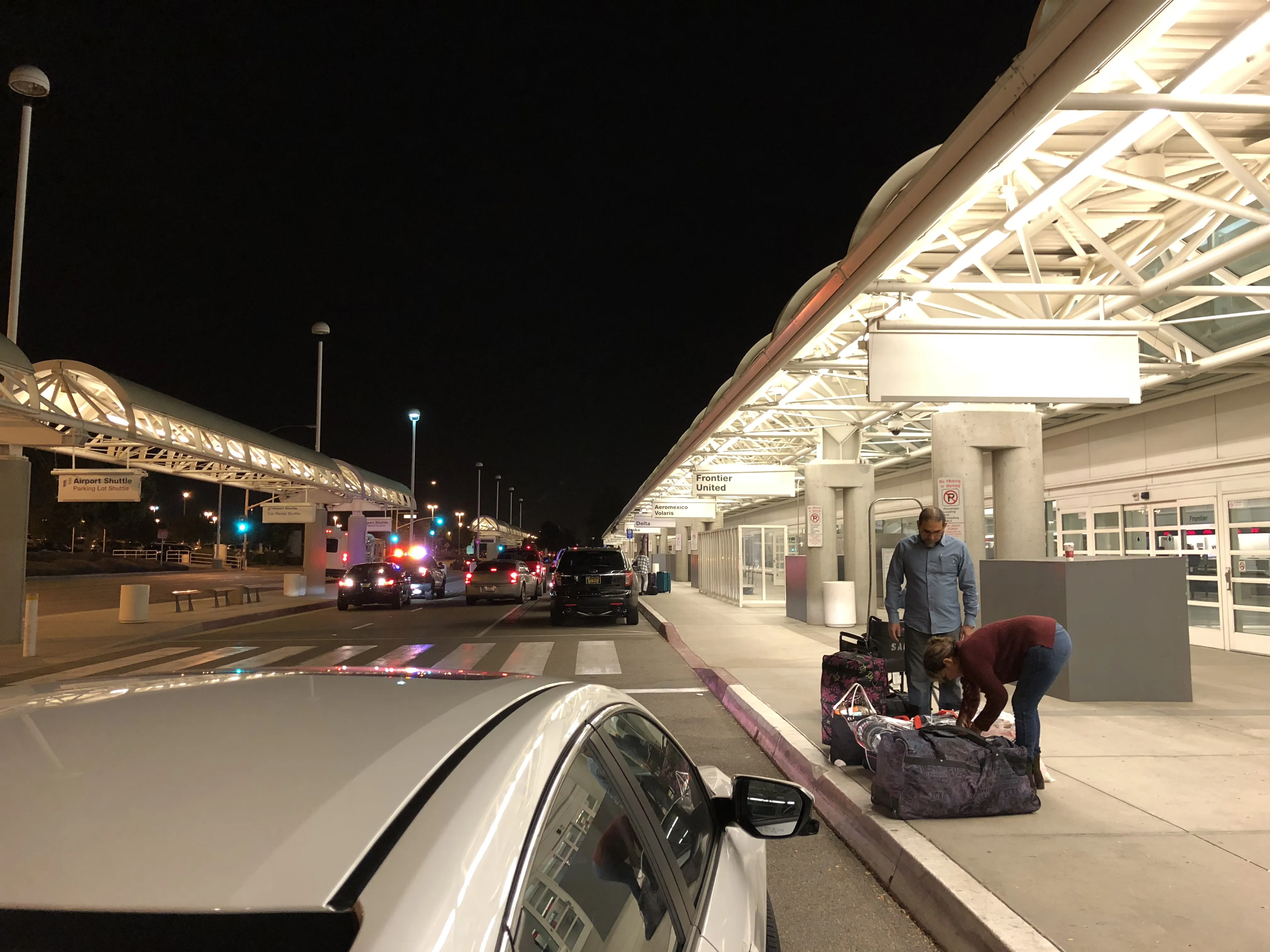 A night view of a brightly lit airport terminal curbside. Travelers are seen loading large suitcases onto the sidewalk under a modern, arched white canopy. Signs for various airlines and airport shuttles are visible above the lanes where several cars are parked or idling against the dark night sky.