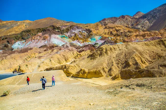 A group of people walks toward hills with vibrant turquoise, pink, and purple mineral deposits, set against tan mountains under a bright blue sky.