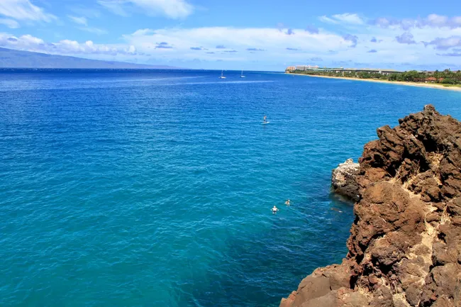 Clear blue water at Maui’s Black Rock with swimmers near the rocky cliff and paddleboarders offshore on a sunny day.