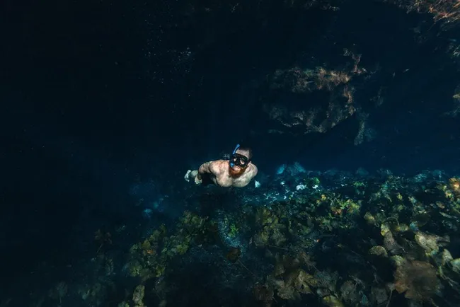 Snorkeler swimming through a dark underwater cavern above rocks and coral.