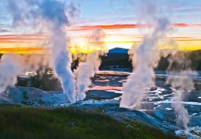 A wide landscape at sunset featuring multiple tall plumes of white steam rising from a geothermal field. The sun hangs low on the horizon behind a dark mountain silhouette, casting vibrant orange, yellow, and red streaks across the sky, which are reflected in the small pools of water on the ground.