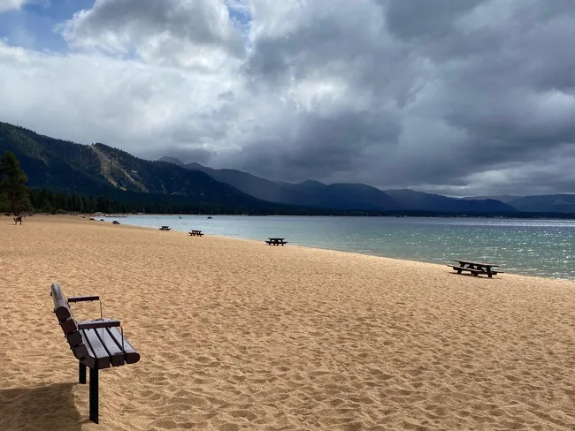 Empty sandy beach with benches and picnic tables beside a calm lake, backed by forested mountains under cloudy skies.