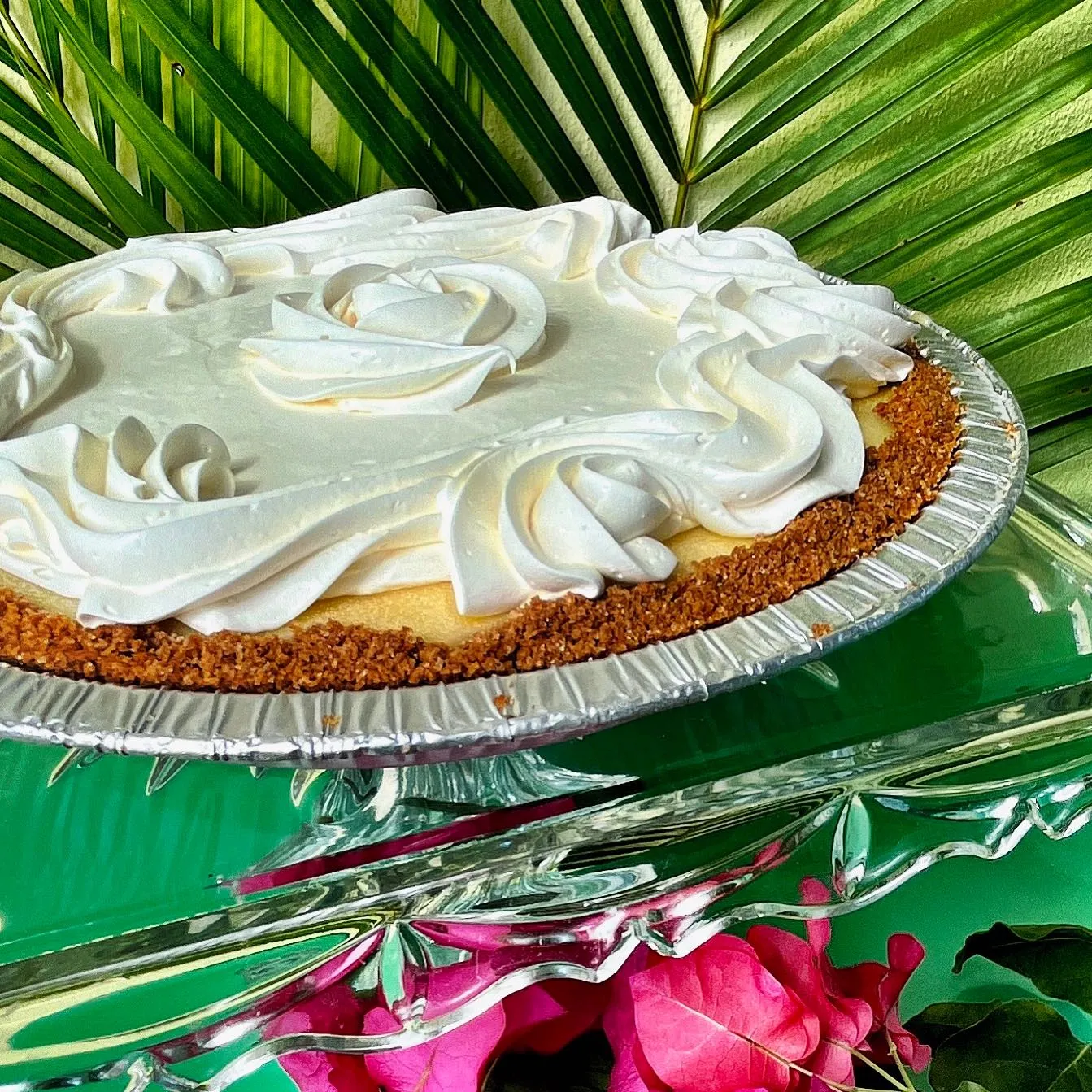 A side-angle, close-up shot of a full key lime pie in a silver foil pan, resting on a decorative glass cake stand. The pie features a thick, textured Graham cracker crust and is topped with smooth, white whipped cream piped into large, elegant rosettes around the edges and a single rose shape in the center. The background is a vibrant green palm frond with bright pink bougainvillea flowers at the bottom, creating a tropical summer feel.