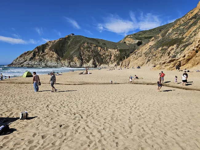 A wide sandy beach sits beneath rugged coastal cliffs, with visitors relaxing, walking, and playing along the shoreline on a sunny day. Gentle waves roll in from the ocean while dramatic rock formations and green-covered hills rise behind the cove under a bright blue sky.