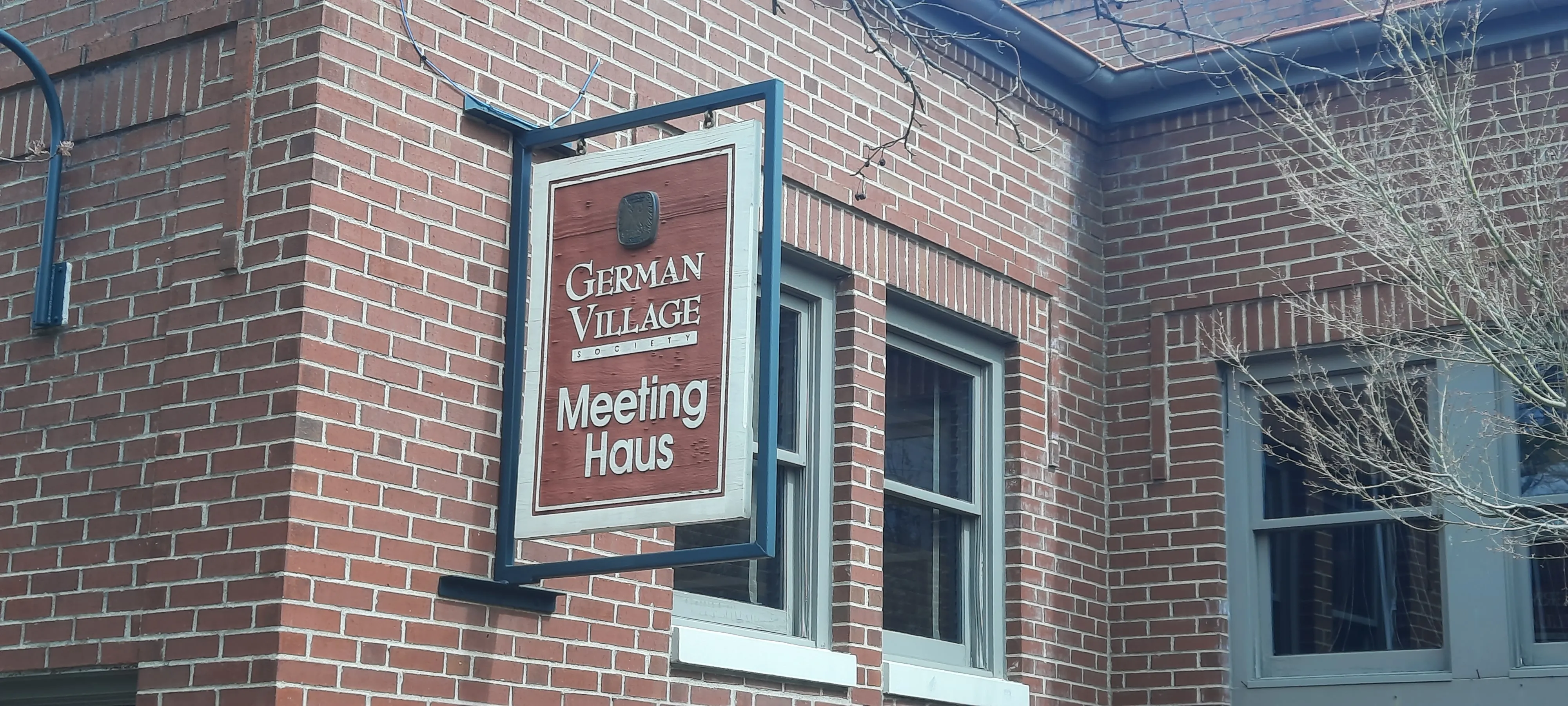 A close-up of a projecting sign mounted on the exterior brick wall of a building. The sign has a dark blue metal frame and a red-brown wooden background with white text reading "German Village Society — Meeting Haus," topped with a small emblem or crest. The building behind it is red brick with gray-trimmed windows. Bare tree branches are visible in the upper right corner.