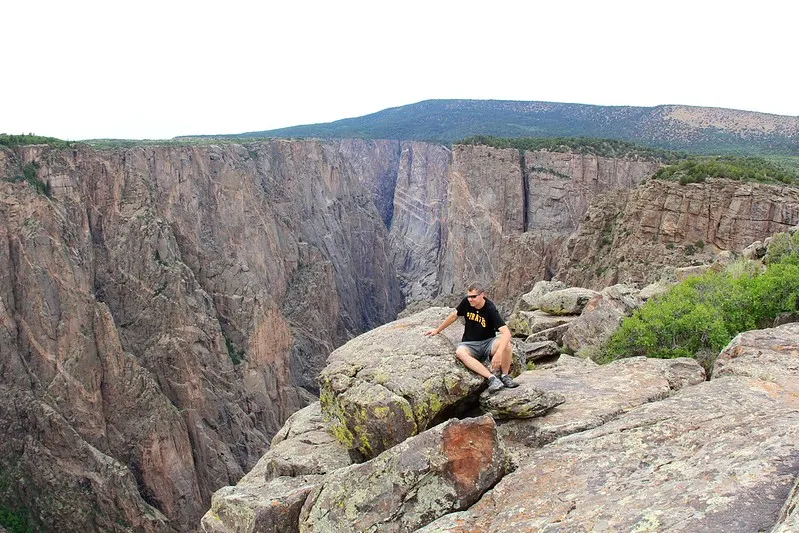 A hiker sits on a rocky ledge overlooking the massive vertical cliffs and deep gorge of Black Canyon under an overcast sky.