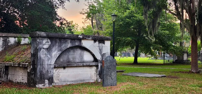 A weathered mausoleum and grave marker in Colonial Park Cemetery, surrounded by lush greenery and a lamp post.