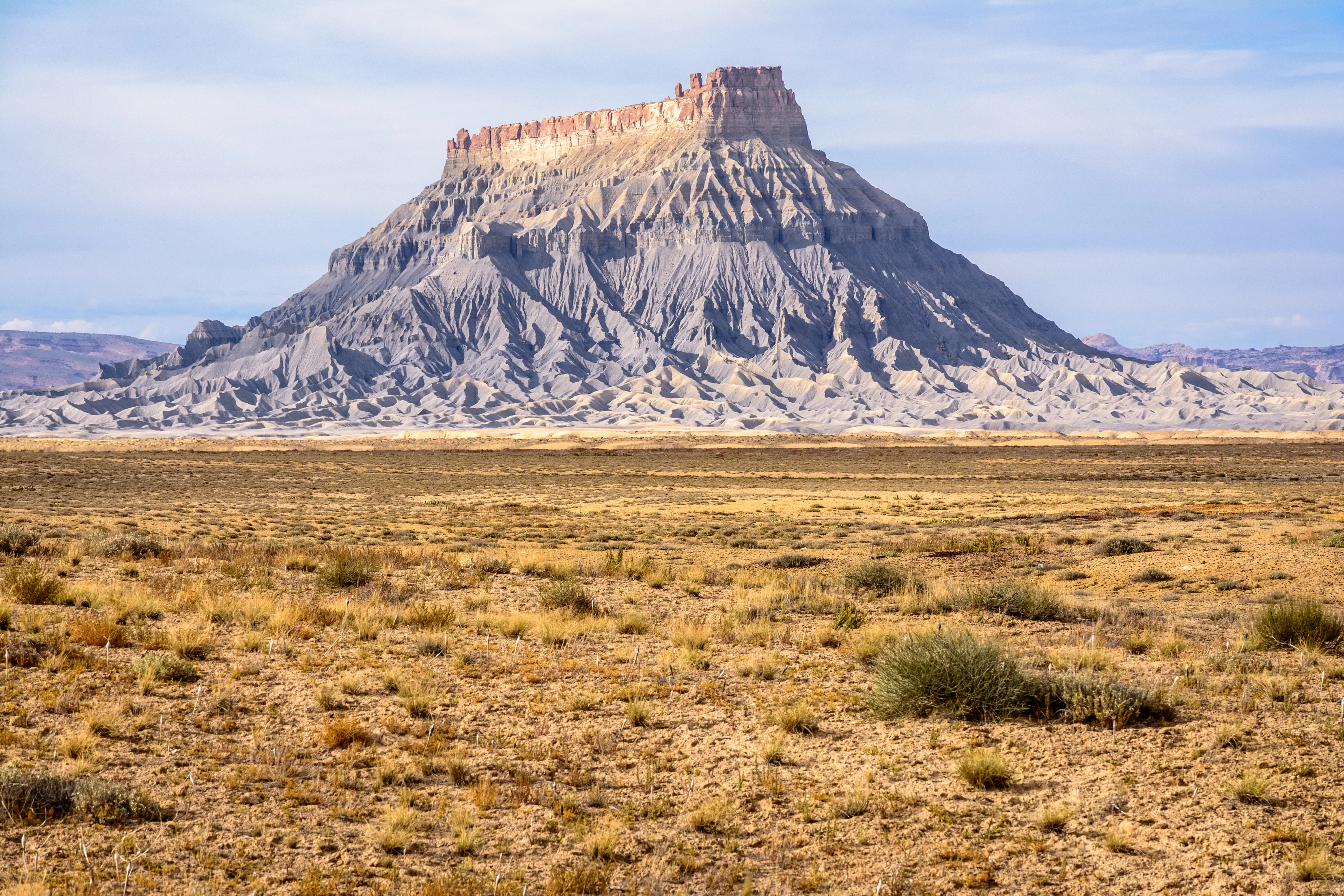 A wide-angle view of Factory Butte, a dramatic flat-topped mesa near Capitol Reef National Park in Utah. The imposing landform rises steeply from a vast, arid desert plain covered in sparse dry grasses and sagebrush. Its lower flanks are composed of deeply eroded gray and tan badlands formations, topped by a cap of reddish-orange sandstone. The scene is photographed under a partly cloudy pale blue sky.