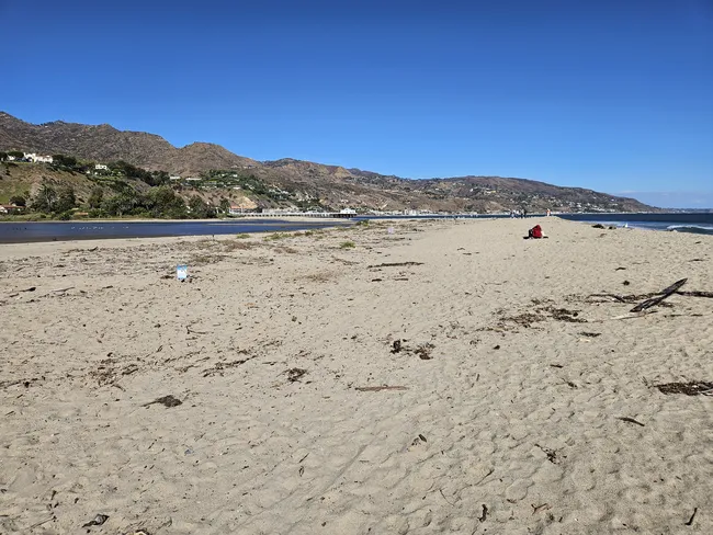 A wide-angle view of a broad sandy beach that acts as a natural barrier between a calm lagoon on the left and the ocean on the right. In the background, rugged brown coastal mountains are dotted with white houses under a clear blue sky. A few distant figures and the white structure of a pier are visible where the sand meets the water in the center of the frame.