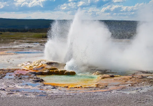 A powerful geyser erupts, sending a tall, wide plume of white water and steam high into the air from a rocky mineral vent. In the foreground, a shallow geothermal pool shows vibrant shades of turquoise, yellow, and orange along its edges, set against a vast, flat landscape under a bright, cloudy sky.