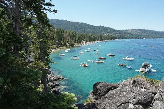 Boats floating on the clear turquoise water of Lake Tahoe near a forested shoreline and rocky cliffs.