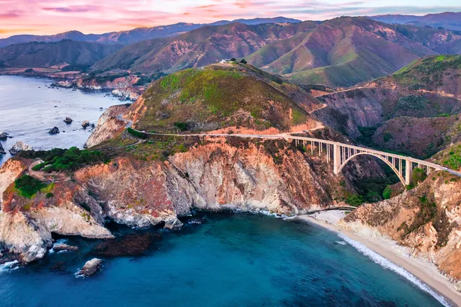 Bixby Creek Bridge spans a rugged coastal canyon along California’s Big Sur coastline, overlooking turquoise ocean waters, rocky cliffs, and rolling green hills beneath a colorful sunset sky.