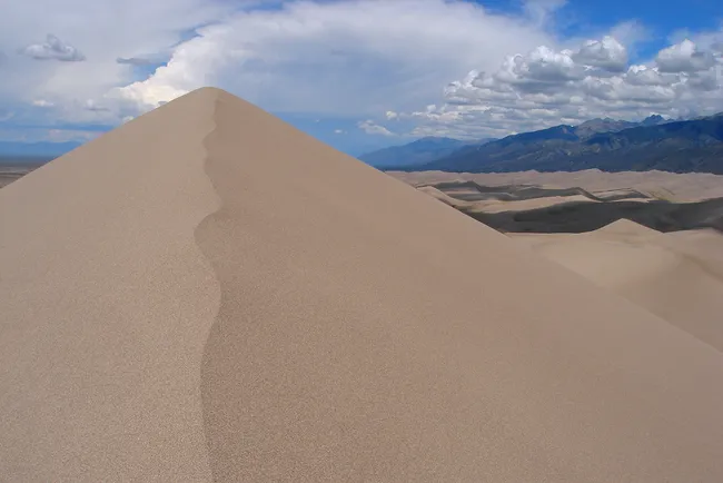 A sharp ridge of Star Dune rises above the vast sandy landscape of Great Sand Dunes National Park, with distant mountains and a cloudy blue sky in the background.