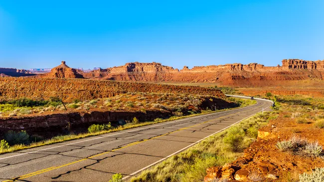A winding two-lane highway — U.S. Route 163 near Mexican Hat, Utah — curves through a dramatic red rock desert landscape on the northern edge of the Navajo Nation. Vivid orange and rust-colored sandstone mesas, buttes, and cliffs dominate the background, with the distant silhouettes of Monument Valley's iconic formations faintly visible on the far left horizon. Sparse desert scrub and sagebrush line the roadside, and a single vehicle travels along the empty road under a cloudless, deep blue sky.