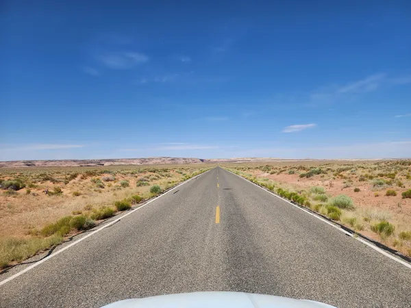 A long, empty two-lane road stretches through flat desert terrain under a cloudless blue sky.