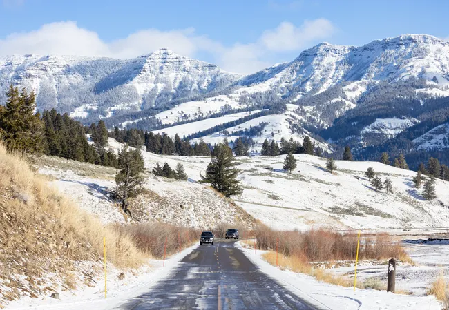 A narrow road cuts through a snowy valley as two vehicles drive toward distant, sunlit mountains covered in white.