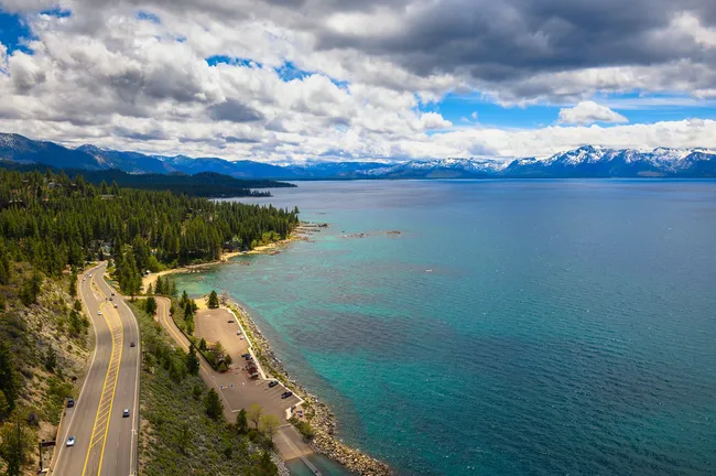Scenic aerial view of a winding lakeside road beside turquoise water, dense pine forest, and distant snow-capped mountains.