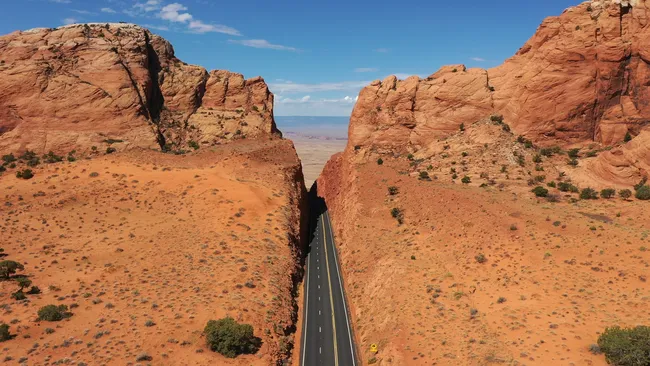 A striking high-angle, vertical aerial shot of a two-lane asphalt highway passing through a narrow, man-made cut in a massive red rock formation. The steep, jagged orange-red cliffs tower over the thin black road, which stretches straight toward a vast, hazy desert horizon in the distance. The surrounding landscape is a mix of red sand and sparse green desert shrubs under a bright, clear blue sky.