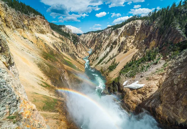 A powerful waterfall plunges into a deep canyon, creating mist and a vivid rainbow above a winding river between steep, colorful cliffs under a bright blue sky.