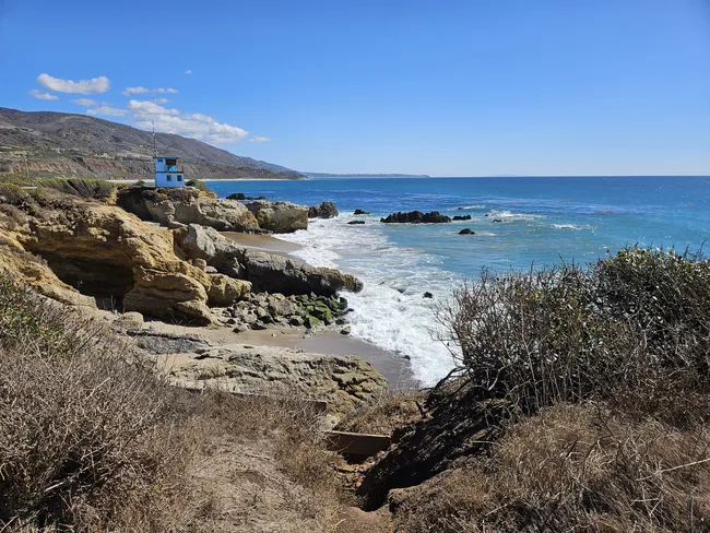 A high-angle view of a rocky coastline where golden-brown bluffs meet a vibrant blue ocean. White waves crash against scattered sea rocks and a small sandy cove. On a prominent rocky outcrop in the mid-ground stands a small, bright blue lifeguard tower, with rolling coastal hills stretching into the distance under a clear sky.