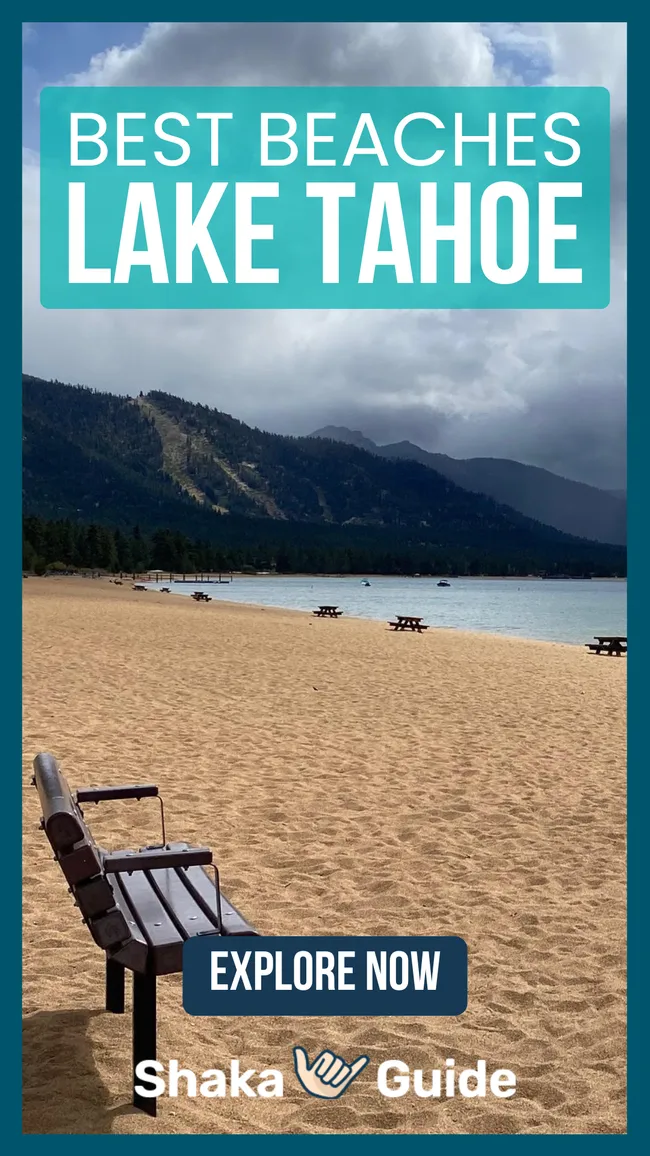 Sandy beach with benches and picnic tables beside a calm lake, surrounded by forested mountains under cloudy skies.