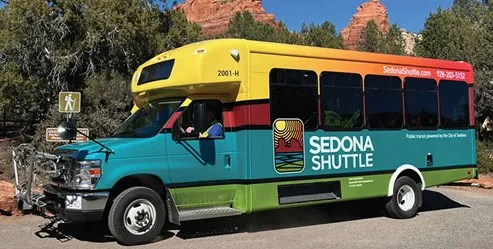 A colorful Sedona Shuttle bus parked near red rock formations, used for transportation to hiking trailheads.
