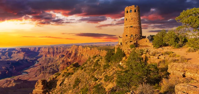 A horizontal, wide-angle landscape shot of the Desert View Watchtower at sunset. The 70-foot-tall stone tower stands on a rocky promontory overlooking the vast, deep expanses of the Grand Canyon. The sky is a dramatic mix of fiery orange and purple clouds. In the distance, the Colorado River winds through the canyon floor. The tower features irregular stone masonry and small, scattered windows, blending seamlessly into the rugged, sun-drenched canyon walls and sparse green desert trees in the foreground.