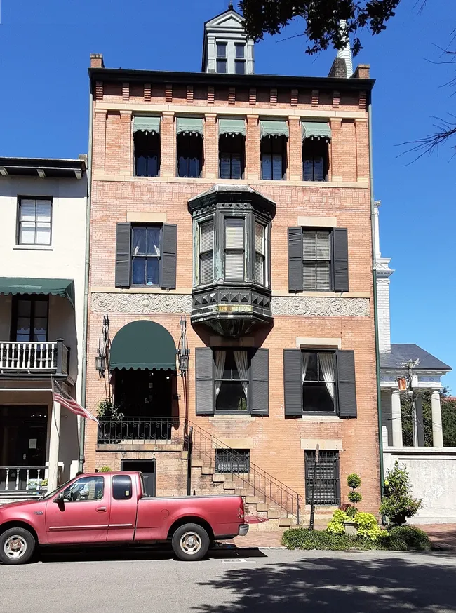 Historic red brick building with a prominent bay window, surrounded by greenery and a parked red pickup truck.