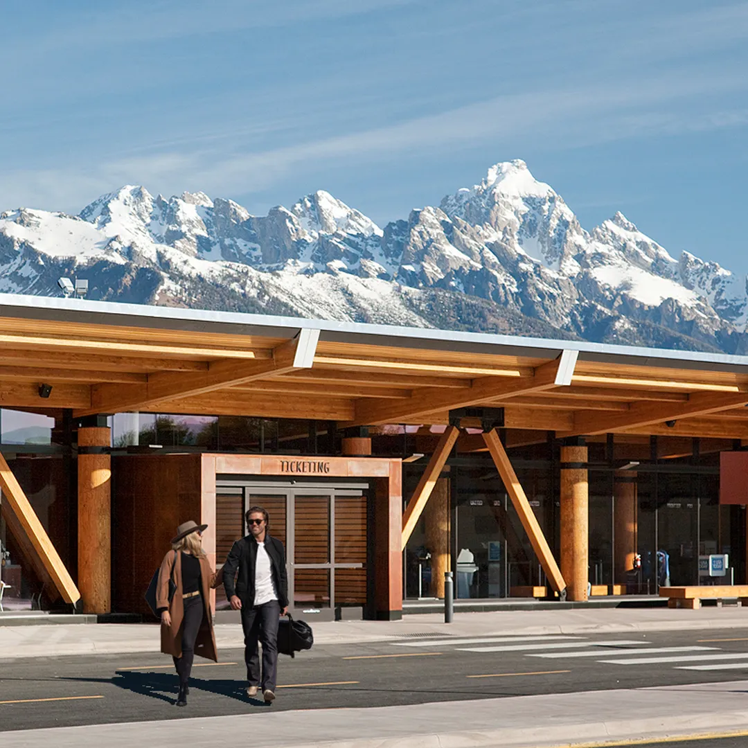 Two travelers walk outside Jackson Hole Airport with snow-covered Teton Range in the background.