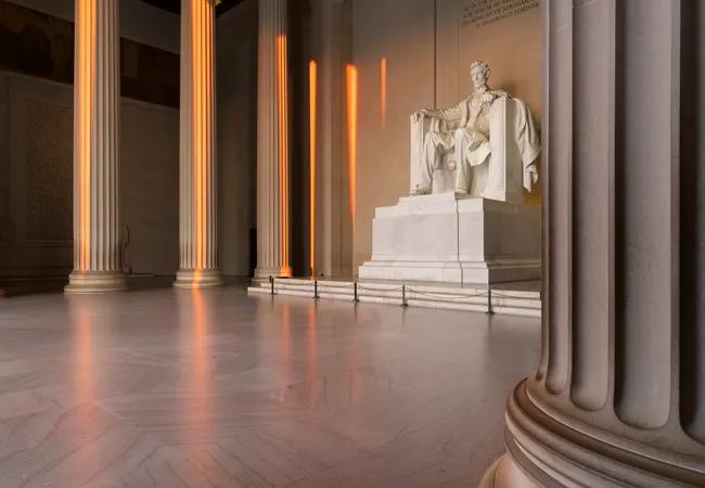 Interior of the Lincoln Memorial in Washington, D.C., photographed at sunrise. Warm golden light streams through the tall Doric columns, casting glowing orange reflections on the polished marble floor. The iconic seated white marble statue of Abraham Lincoln is centered in the background, bathed in soft morning light. The grand, serene atmosphere of the empty chamber emphasizes the memorial's majestic scale and quiet dignity.