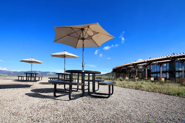 Picnic tables with umbrellas sit outside the Great Sand Dunes Visitor Center under a clear blue sky.