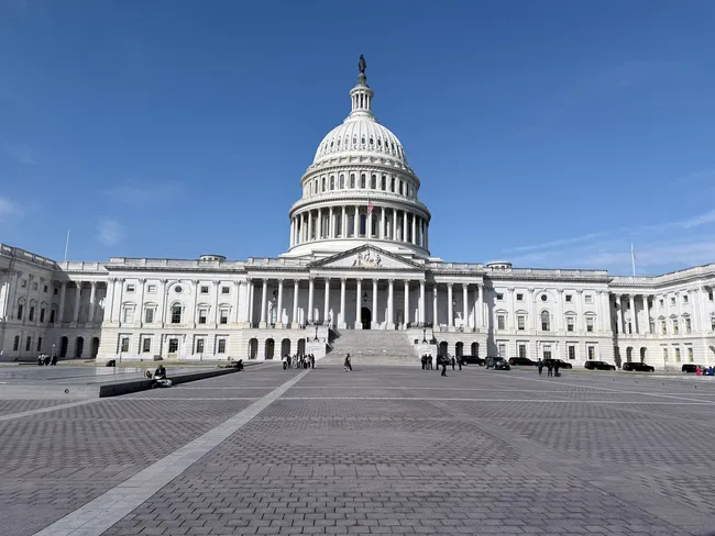 A wide-angle view of the United States Capitol building in Washington, D.C., photographed from the East Plaza on a clear, sunny day. The iconic white neoclassical structure features a large central dome topped with the Statue of Freedom, flanked by symmetrical colonnaded wings. A broad brick-paved plaza stretches across the foreground, with a small number of visitors and parked vehicles visible near the building's entrance steps. The scene is set against a deep, cloudless blue sky.