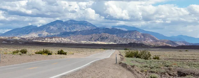 Long desert highway stretching toward distant mountains under a partly cloudy sky with sparse vegetation along the roadside.