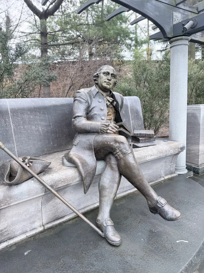 A bronze statue of Benjamin Franklin seated casually on a granite bench, depicted in 18th-century attire — knee breeches, buckled shoes, a long coat, and ruffled shirt — wearing glasses and holding a document or book. A stack of books rests beside him on the bench, and a walking cane leans against it. An inscription is carved into the stone wall behind the figure. A metal pergola structure and bare winter trees are visible in the background. The approachable, life-size sculpture invites visitors to sit alongside the figure.