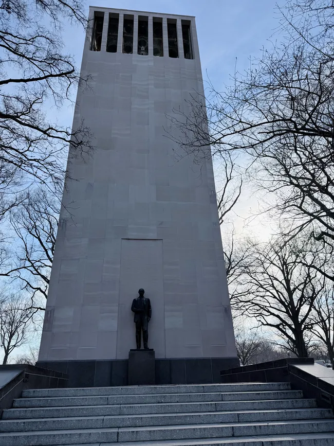 A low-angle view looking up at the Robert A. Taft Memorial and Carillon in Washington, D.C. A tall, stark white marble bell tower rises dramatically against a pale blue sky, with open belfry openings visible at the top housing the carillon bells. A bronze statue of Senator Robert A. Taft stands on a small pedestal at the base of the tower, silhouetted against the light stone. Wide granite steps lead up to the monument, flanked by bare winter trees on both sides.
