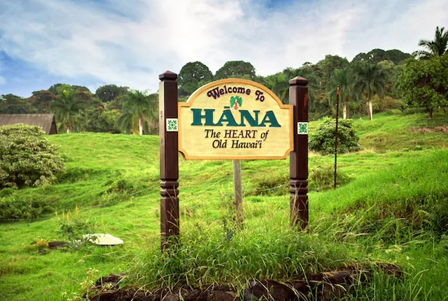 A roadside sign reading “Welcome to Hāna, The Heart of Old Hawaiʻi,” set in a lush green hillside with palm trees.