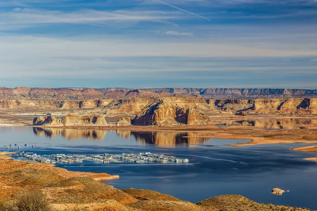 A high-angle, panoramic landscape of Lake Powell featuring Wahweap Marina. In the mid-ground, a large cluster of white boats is docked at a marina in the deep blue water. Behind it, massive red and tan sandstone mesas rise from the lake, creating sharp reflections on the calm surface. The background is a wide, desert plateau stretching to the horizon under a soft blue sky with thin, wispy clouds. The foreground consists of rolling, arid hills covered in sparse desert scrub.
