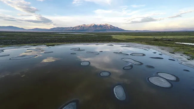 Aerial view of a shallow wetland in Great Sand Dunes National Park with scattered circular patches, reflecting clouds and a distant mountain range at sunset.
