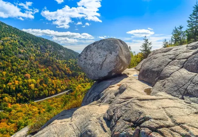Large balancing boulder atop a rocky ledge overlooking Acadia National Park’s colorful autumn forest and mountain valley.