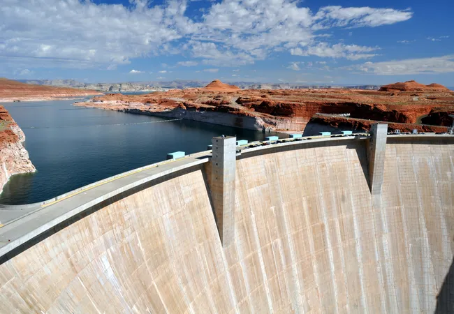A massive curved concrete dam drops steeply toward a river below, with calm blue water and red desert cliffs stretching out behind it.