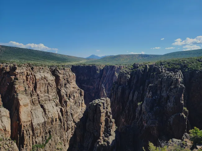 Towering cliffs of Black Canyon of the Gunnison rise sharply on both sides of a deep, narrow gorge, with distant mountain peaks visible under a clear blue sky.