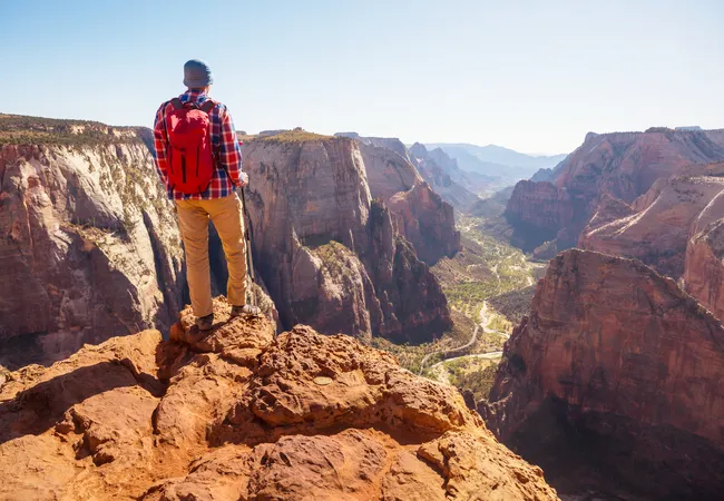 Man with a red backpack stands on a rocky cliff edge, looking out over a deep canyon with towering red cliffs and a winding valley below.