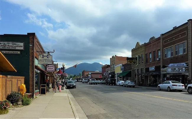 A view of downtown Red Lodge, Montana, with historic brick buildings lining the street, small shops and cafés on the left, parked cars along the road, and forested mountains visible in the distance under a partly cloudy sky.