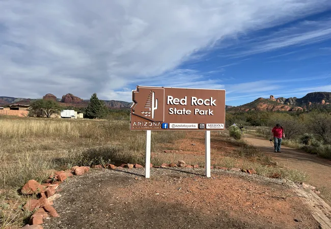 A brown Arizona state park sign marks the entrance to Red Rock State Park with a visitor walking nearby.