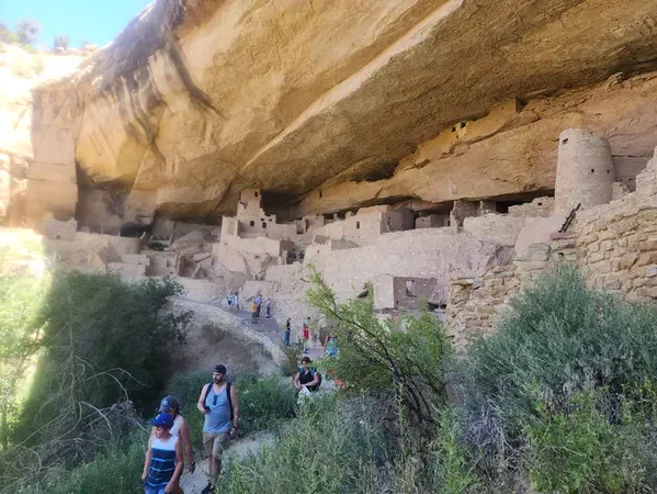 ChatGPT said:  Visitors walking along a trail below massive sandstone cliffs, exploring ancient cliff dwellings with stone towers and rooms built into the rock face.