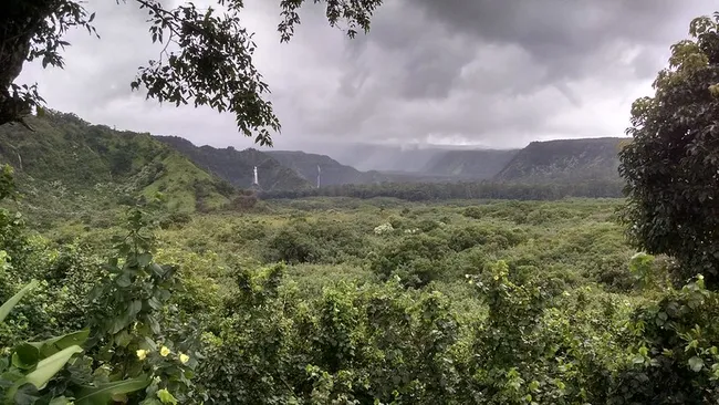 A wide, lush green valley under cloudy skies with twin waterfalls cascading down distant cliffs.